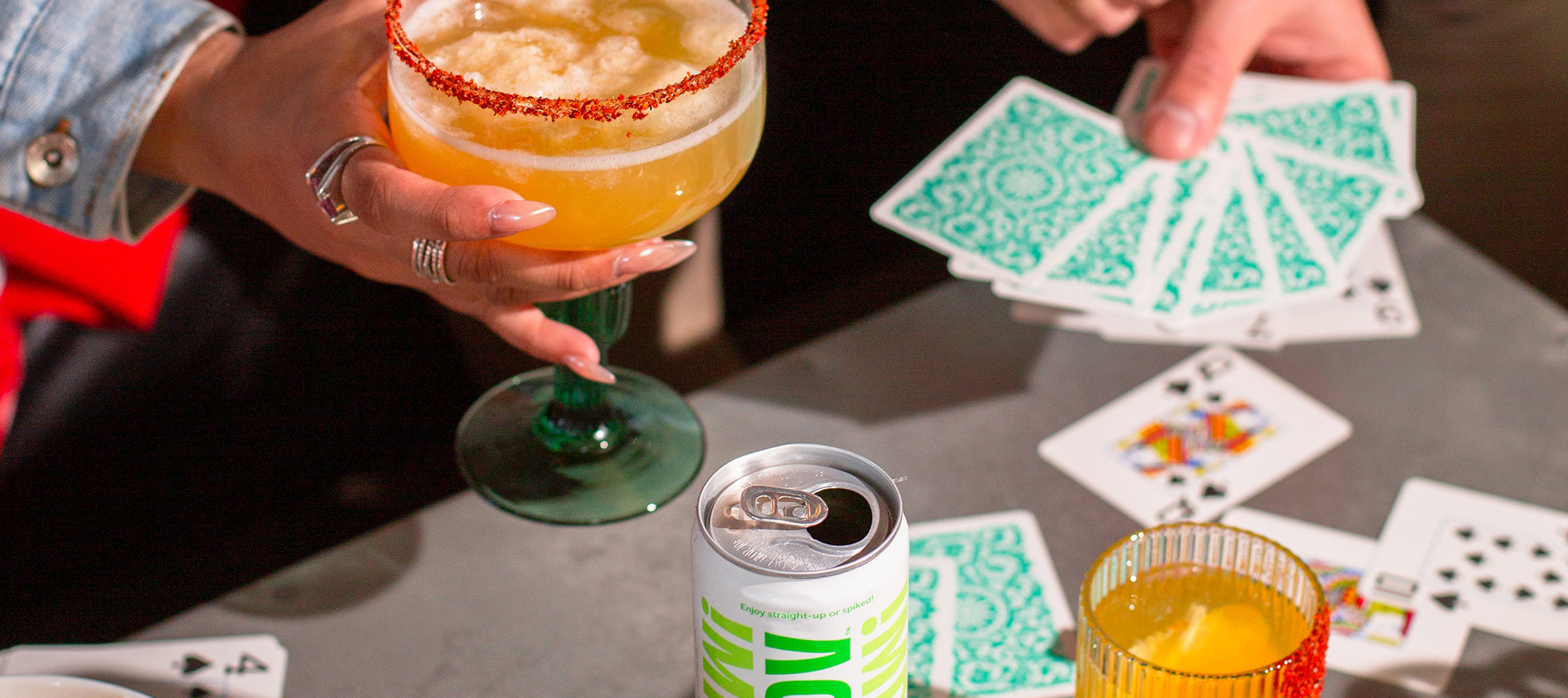 Close-up of people playing cards on grey table with various non-alcoholic cocktails.