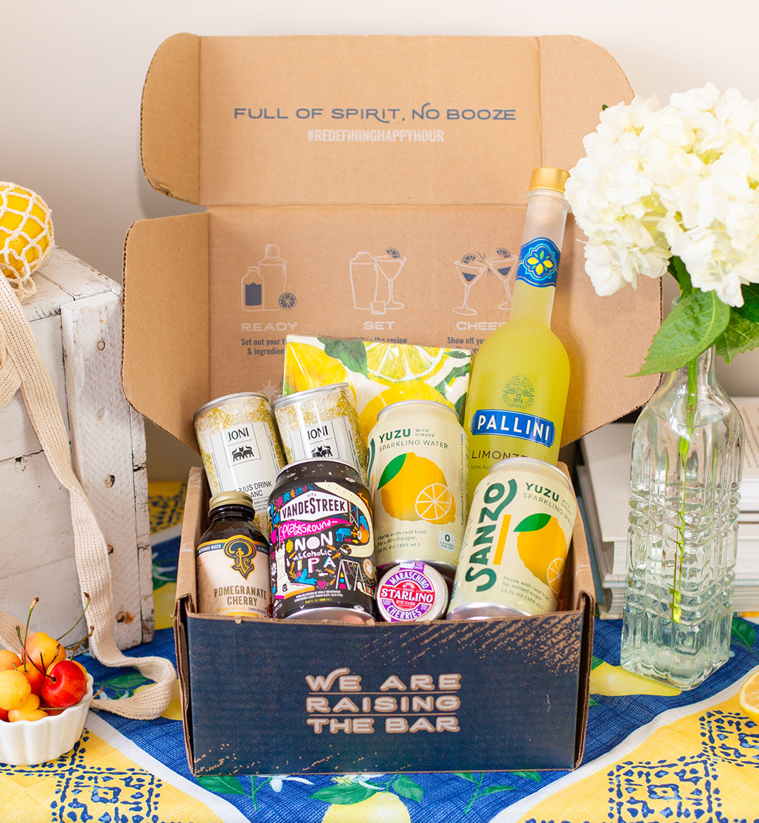 Raising the Bar shipping box with various non-alcoholic drinks inside. Flowers and wooden box on patterned blue and yellow table cloth.