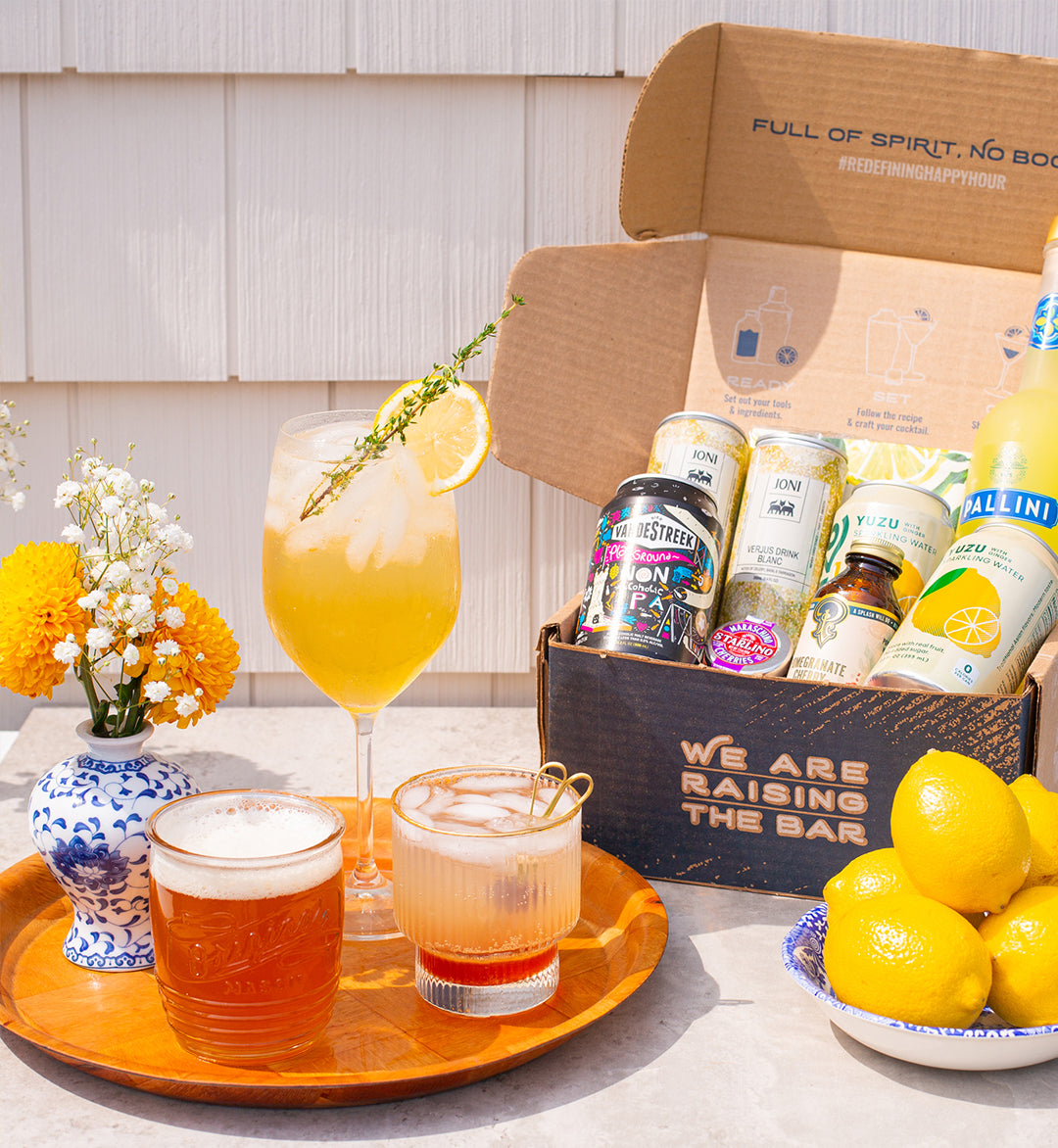 Non-alcoholic cocktails on serving tray with flowers, bowl of lemons and Raising the Bar shipping box with various non-alcoholic drinks inside. Scene is outside with house siding in background.