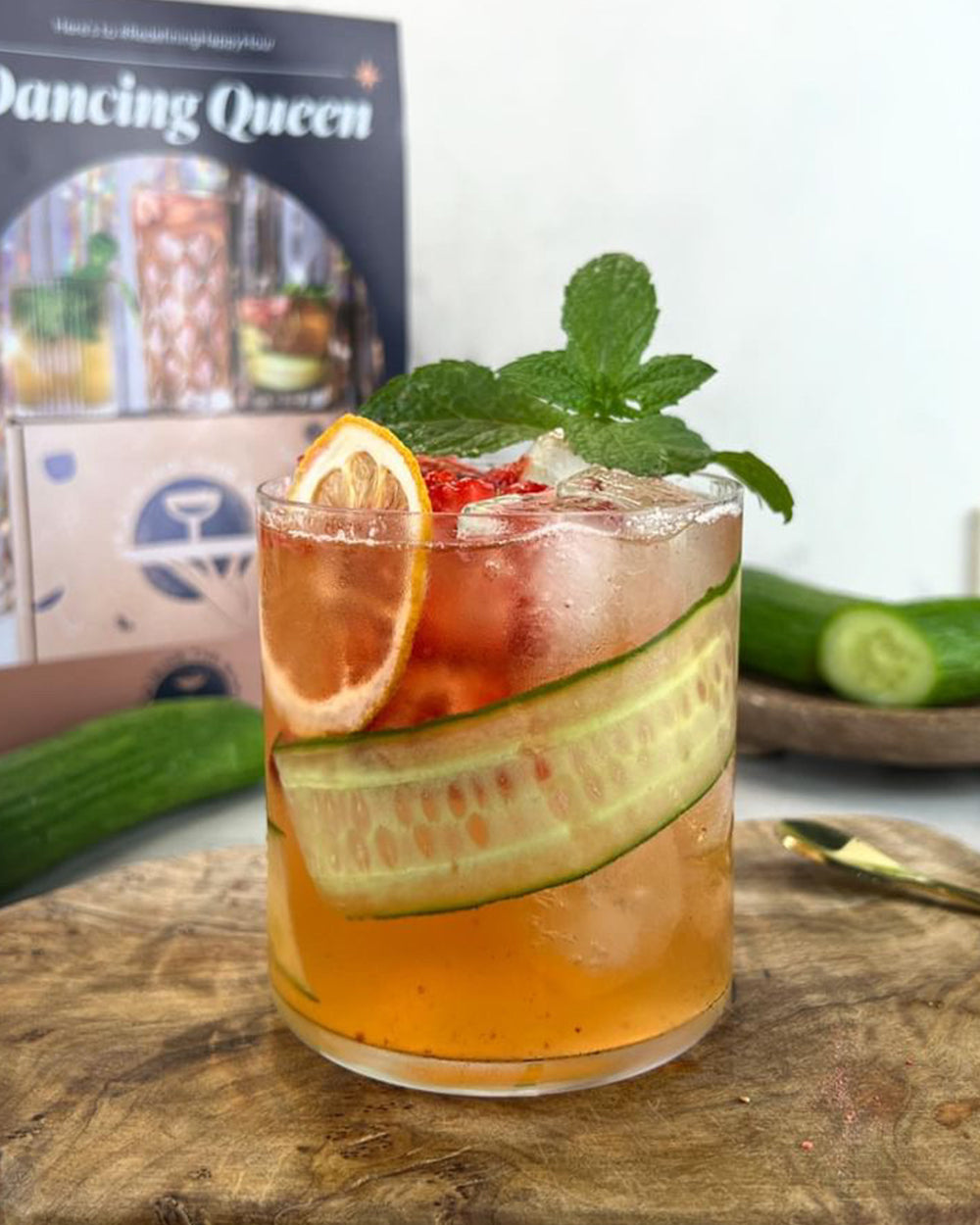 Non-alcoholic cocktail with mint, cucumber and orange garnishes on a wooden cutting board. Cucumbers and box in background.