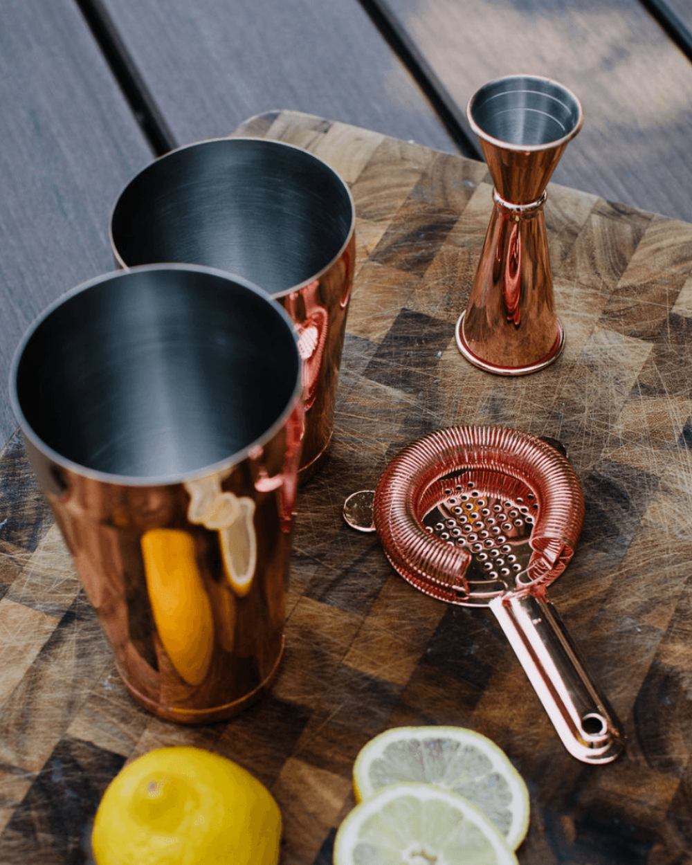 Rose gold cocktail shakers and strainer on a wooden surface with lemons and limes.