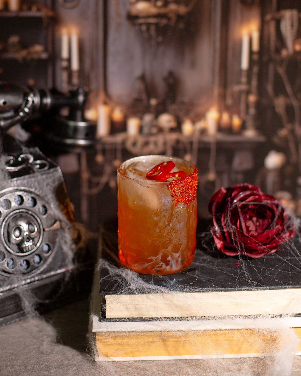 Fire & Brimstone zero-proof cocktail on stack of books with cobwebs, wilted rose and old phone on table.