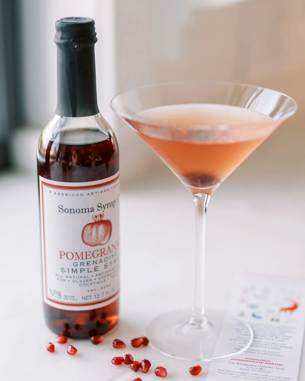 Bottle of Sonoma Syrup with a pink cocktail and pomegranate seeds on a white surface