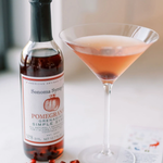 Bottle of Sonoma Syrup with a pink cocktail and pomegranate seeds on a white surface
