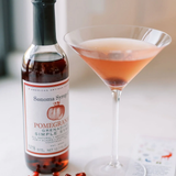 Bottle of Sonoma Syrup with a pink cocktail and pomegranate seeds on a white surface