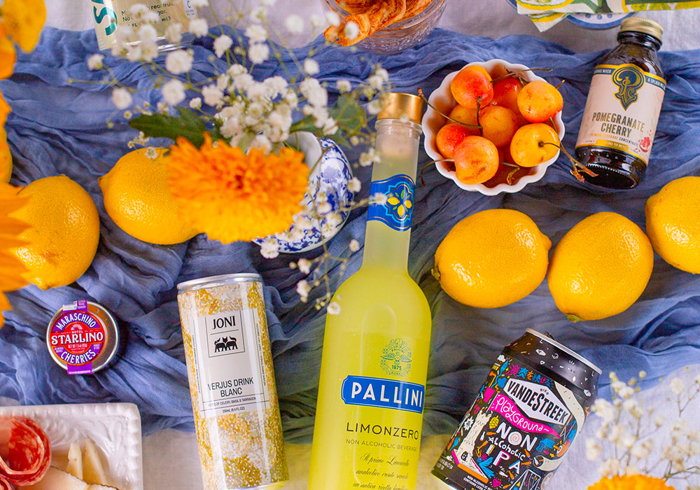 Various non-alcoholic cocktails and ingredients scattered on table with various fruits, flowers and blue cloth.