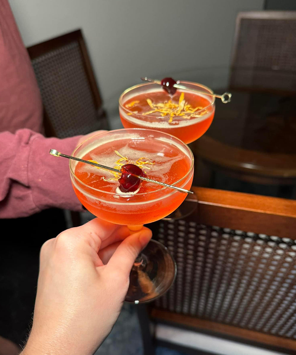 Two people cheersing orange non-alcoholic cocktails with glass table in background.
