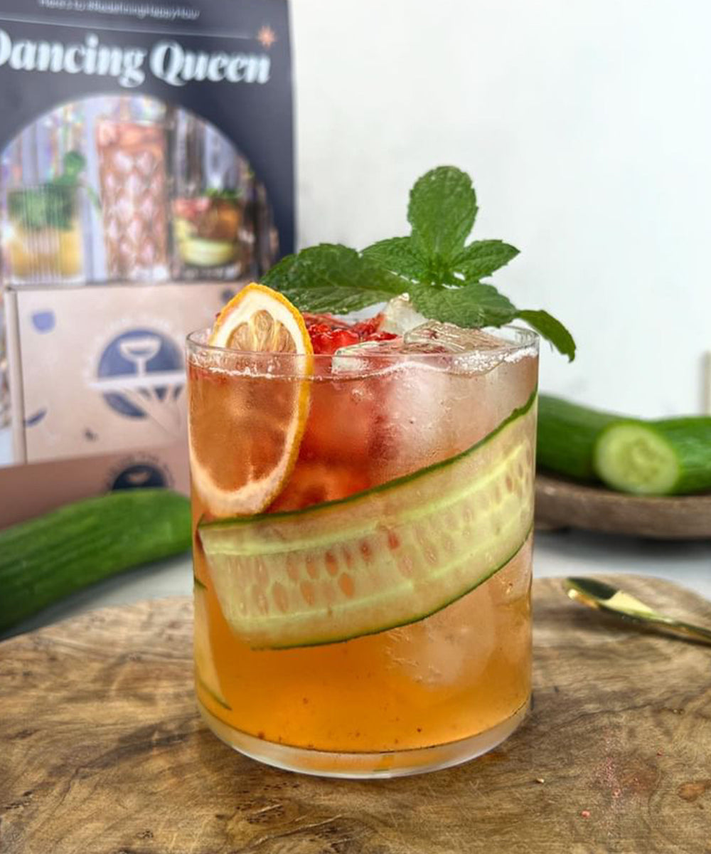 Non-alcoholic cocktail with mint, cucumber and orange garnishes on a wooden cutting board. Cucumbers and box in background.