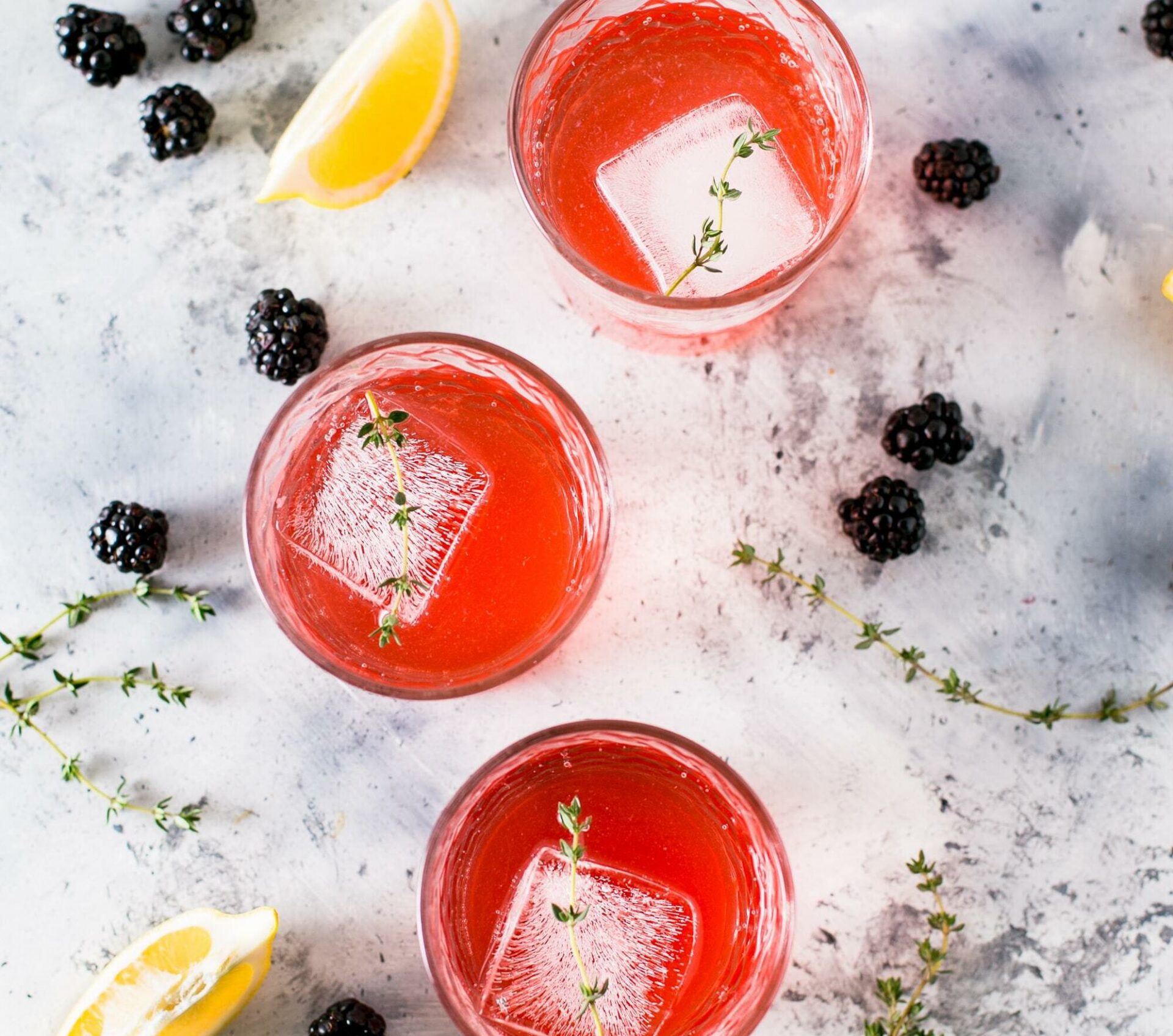 Three glasses of alcohol-free blackberry lemonade with ice and thyme.