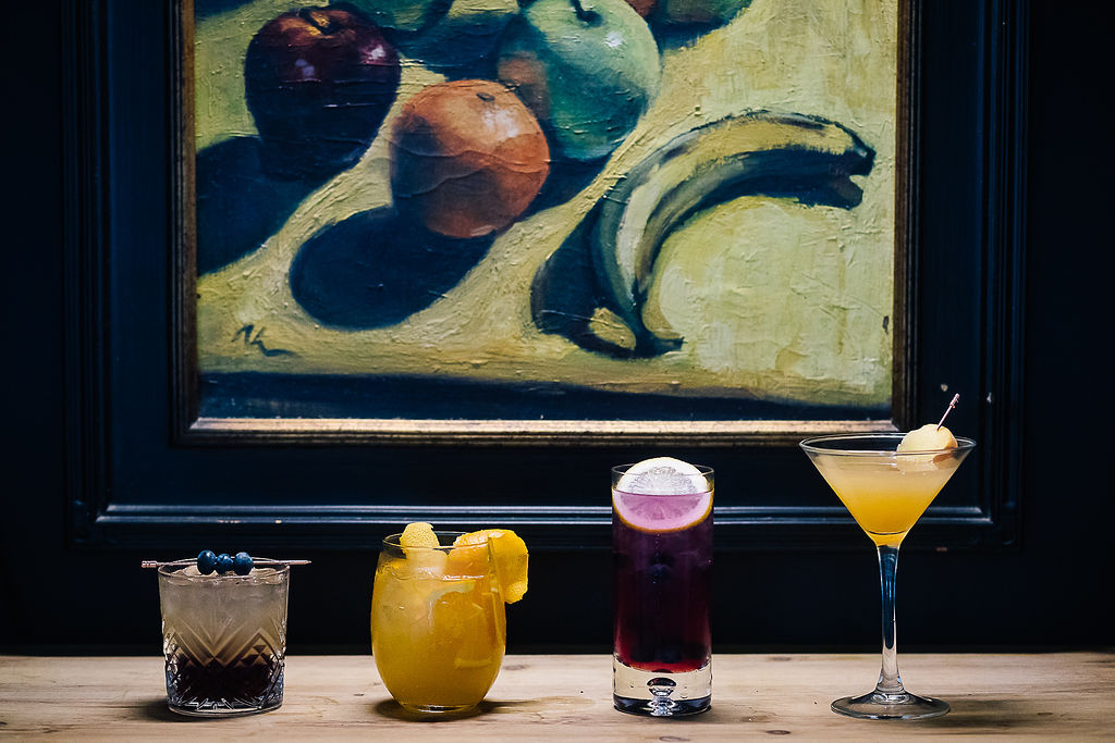 Three cocktail drinks on a table next to a painting during happy hour.